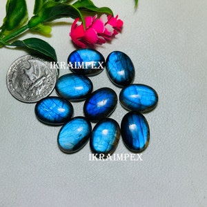 May include: A collection of eight oval-shaped, blue labradorite gemstones with iridescent flashes of color. The stones are arranged in a cluster on a light gray surface. The text "IKRAIMPEX" appears twice in the image.