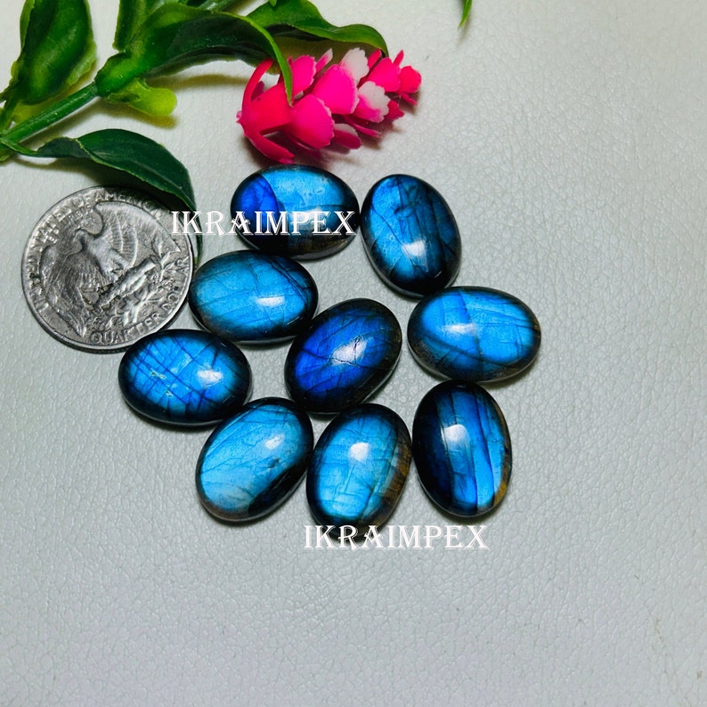 May include: A collection of eight oval-shaped, blue labradorite gemstones with iridescent flashes of color. The stones are arranged in a cluster on a light gray surface. The text "IKRAIMPEX" appears twice in the image.