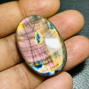 K&ouml;nnte beinhalten: Ovaler Labradorit-Cabochon mit irisierenden blauen, gr&uuml;nen und goldenen Schimmern. Der Stein hat eine rosa-graue Grundfarbe.