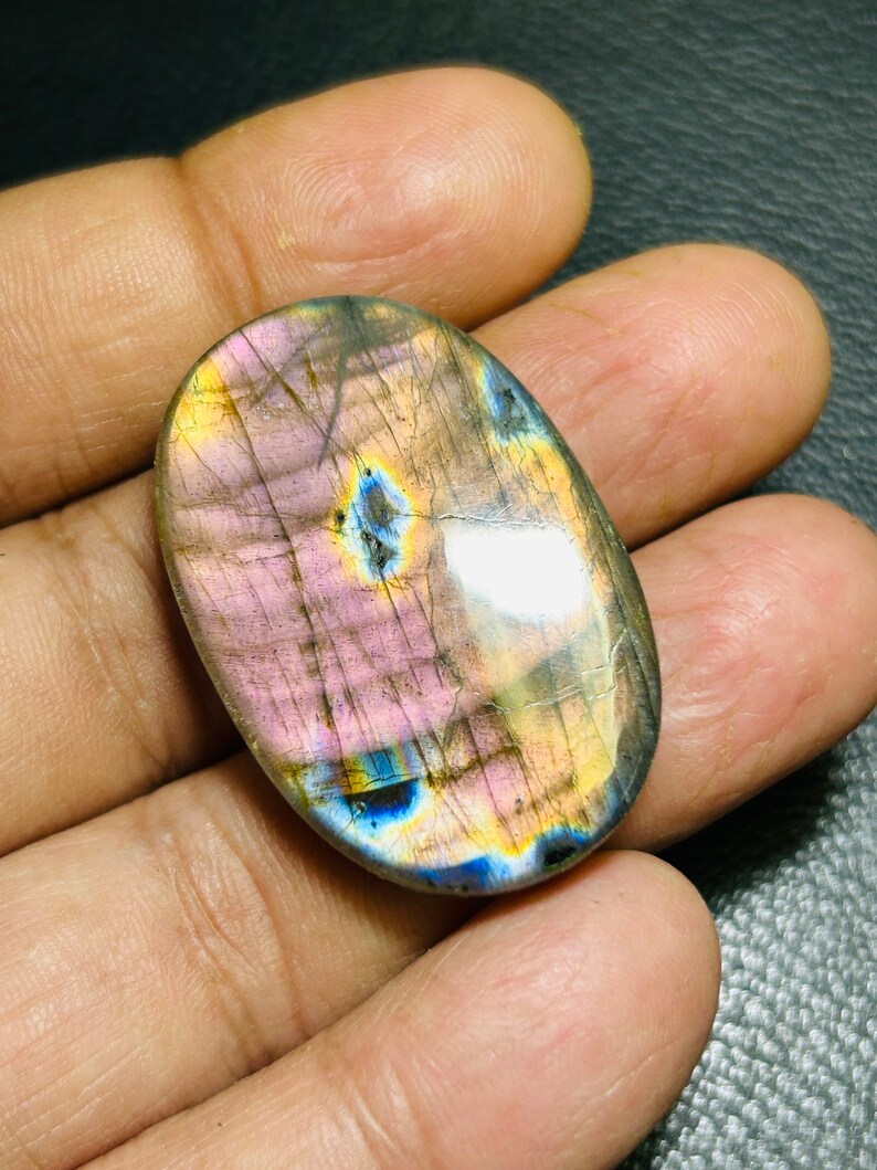 K&ouml;nnte beinhalten: Ovaler Labradorit-Cabochon mit irisierenden blauen, gr&uuml;nen und goldenen Schimmern. Der Stein hat eine rosa-graue Grundfarbe.
