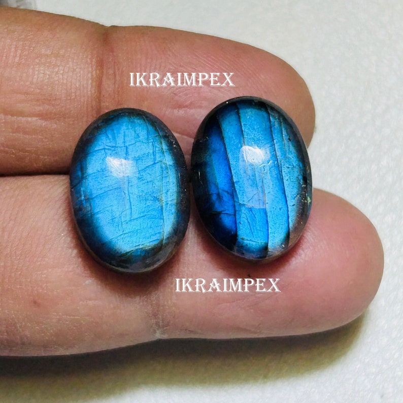 May include: Two oval-shaped blue labradorite gemstones with iridescent flashes of color. The stones are set against a light brown background.