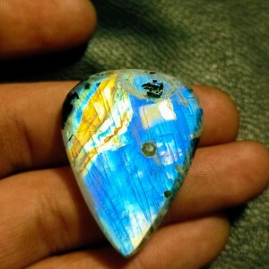 May include: A blue and gold iridescent labradorite gemstone cabochon with a teardrop shape. The gemstone has a unique pattern of blue, gold, and white.