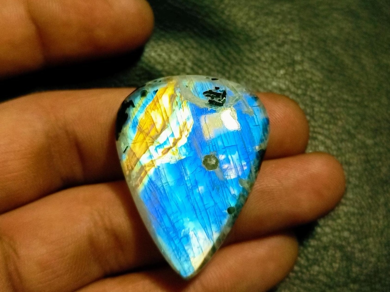 May include: A blue and gold iridescent labradorite gemstone cabochon with a teardrop shape. The gemstone has a unique pattern of blue, gold, and white.