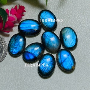 May include: Eight oval-shaped labradorite gemstones with iridescent blue and gray hues. The stones are arranged in a cluster on a gray surface. The text "IKRAIMPEX" appears twice in the image.
