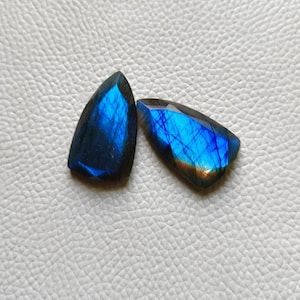 May include: Two polished labradorite gemstones with a striking blue iridescence. The stones have a dark base color with flashes of blue and a hint of orange. The gemstones are cut into unique shapes, perfect for jewelry making.