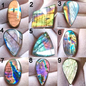 Puede incluir: Una colección de nueve piedras preciosas de labradorita pulidas, cada una con formas únicas y patrones de color iridiscentes. Las piedras muestran una gama de colores, incluyendo azules, verdes, púrpuras y dorados, con variaciones en forma y tamaño. Las piedras preciosas están numeradas del 1 al 9.
