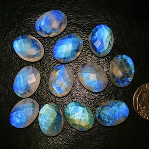 Puede incluir: Una colección de piedras preciosas de piedra lunar de forma ovalada con un corte facetado. Las piedras muestran una gama de colores, incluyendo azules, blancos y toques de amarillo. Las piedras preciosas están dispuestas sobre una superficie oscura.