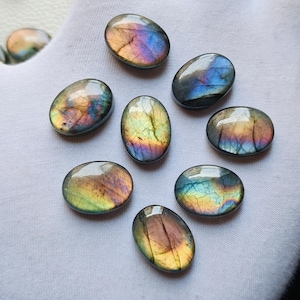 Pode incluir: Oito pedras preciosas ovais de labradorita com flashes iridescentes de azul, roxo, amarelo e laranja. As pedras polidas são exibidas sobre um tecido branco, mostrando suas cores e padrões naturais. Ideais para a criação de joias.