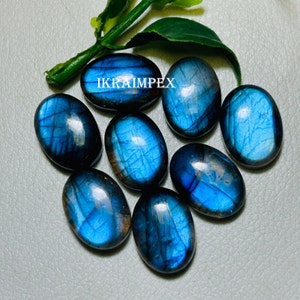 May include: Eight oval-shaped labradorite gemstones with a blue iridescent sheen. The stones are arranged in a cluster on a gray surface. The text "IKRAIMPEX" is visible in the image.