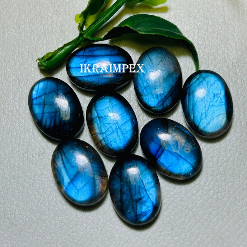 May include: Eight oval-shaped labradorite gemstones with a blue iridescent sheen. The stones are arranged in a cluster on a gray surface. The text "IKRAIMPEX" is visible in the image.