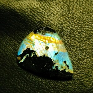 May include: A triangular iridescent labradorite gemstone with a black, blue, and yellow color pattern. The gemstone is set against a dark green leather background.
