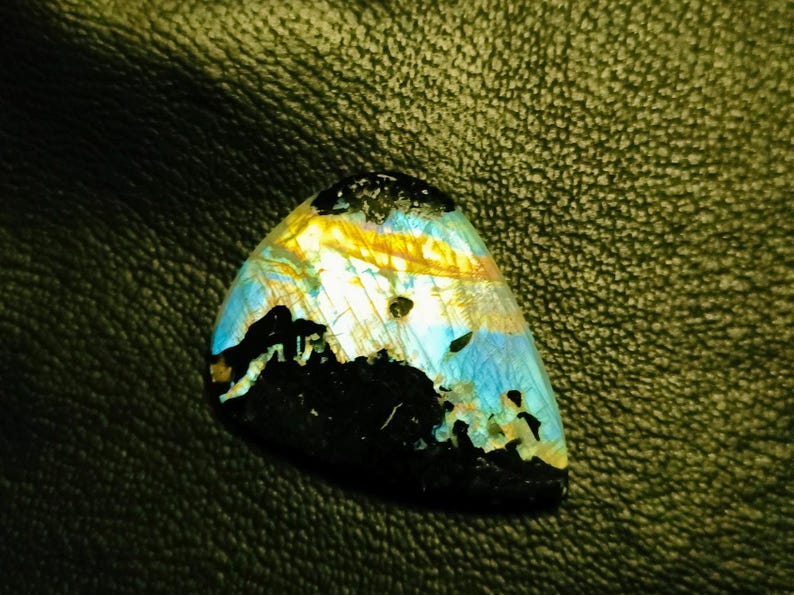 May include: A triangular iridescent labradorite gemstone with a black, blue, and yellow color pattern. The gemstone is set against a dark green leather background.