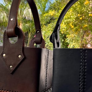 Puede incluir: Dos bolsos tote de cuero. Uno es marrón oscuro con una base cosida y fijaciones de asa aseguradas con remaches. El otro es negro con una costura lateral cosida y un asa. Ambos tienen asas arqueadas.