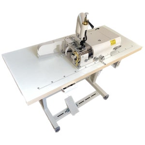 Peut inclure: Machine à coudre industrielle blanche avec une pédale au pied sur une table blanche avec une règle. La machine a un corps blanc et argenté avec une poignée noire.