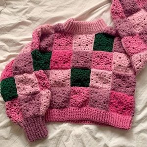 Könnte beinhalten: Ein rosa und grüner gehäkelter Pullover mit einem quadratischen Muster. Der Pullover hat lange Ärmel und einen runden Ausschnitt.