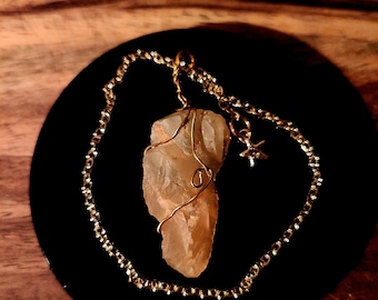 May include: A natural quartz crystal pendant with a gold-colored wire wrap and chain. The crystal is a light amber color and is suspended from a delicate chain with a small star charm. The pendant rests on a black circular surface.