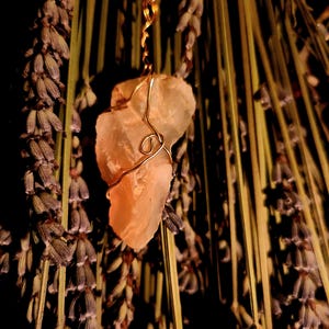May include: A pendant necklace with a raw, pale orange crystal encased in gold-coloured wire. The necklace is set against a backdrop of dried lavender, with a dark background. The crystal is irregularly shaped and has a natural, earthy appearance.
