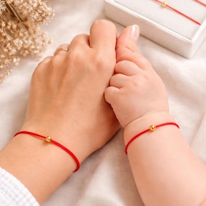 May include: Red string bracelets with gold beads, one on an adult's wrist and one on a baby's wrist. A white box with two similar bracelets is in the background. The bracelets are on a white surface.