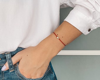 May include: White button-down shirt with a pocket featuring a black line drawing of a woman's silhouette. The shirt has text above the pocket. The person is wearing blue jeans and a red bracelet with a small charm.