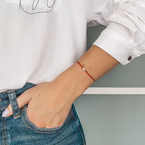 Puede incluir: Camisa blanca abotonada con un bolsillo con un dibujo lineal negro de la silueta de una mujer. La camisa tiene texto encima del bolsillo. La persona lleva vaqueros azules y una pulsera roja con un pequeño dije.