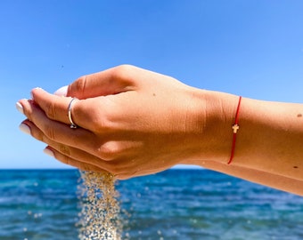 May include: A person's hands releasing sand against a backdrop of a bright blue sky and ocean. A red string bracelet with a small gold cross charm is visible on the wrist. A silver ring is on the finger.