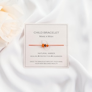 May include: A red string bracelet with three amber beads. The card reads "Child Bracelet Make a Wish" and "Natural Amber Healing Protection Calmness" and "Once the bracelet rips off your hand, your wish becomes reality."