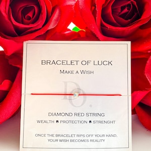 Puede incluir: Una pulsera de hilo rojo con una pequeña gema cuadrada y transparente en el centro. La tarjeta dice "BRACELET OF LUCK" y "MAKE A WISH". También se ve el texto "DIAMOND RED STRING". El fondo presenta rosas rojas.