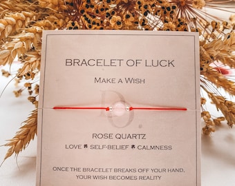 Pode incluir: Um bracelete de corda vermelha com uma conta de quartzo rosa. O cartão diz "Bracelete da sorte, Faça um desejo, Quartzo rosa, Amor, Autoconfiança, Calma, Uma vez que o bracelete se quebre da sua mão, o seu desejo se tornará realidade."