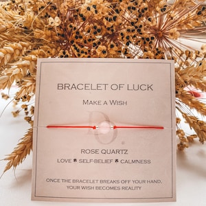 Puede incluir: Un brazalete de cuerda roja con una cuenta de cuarzo rosa. La tarjeta dice "Brazalete de la suerte, Pide un deseo, Cuarzo rosa, Amor, Autoconfianza, Tranquilidad, Una vez que el brazalete se rompa de tu mano, tu deseo se hará realidad."