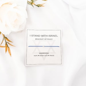 May include: A blue string bracelet with a silver charm in the shape of the Star of David. The bracelet is on a white card with the text "I Stand With Israel Bracelet of Peace" and "Swarovski Hope Good Luck Peace".