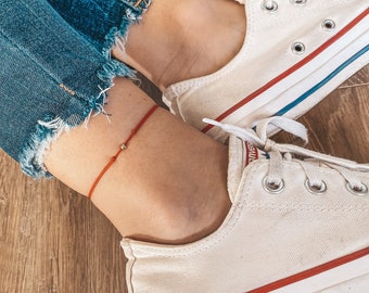 May include: A red string ankle bracelet with a small gold bead charm. The bracelet is worn on a person's ankle with white sneakers.
