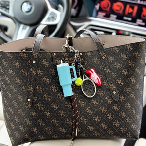 Puede incluir: Un bolso tote marrón con el logotipo de Guess y detalles dorados. El bolso tiene varios abalorios colgados, incluyendo una botella de desinfectante de manos azul, una raqueta de tenis, una pelota de tenis y un abalorio de zapato rojo.