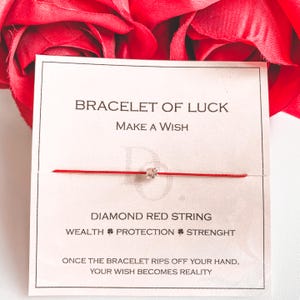 Op de afbeelding: Een rode draadarmband met een kleine diamantachtige bedel. De kaart leest "BRACELET OF LUCK" en "DIAMOND RED STRING". Extra tekst is "WEALTH PROTECTION STRENGHT" en "ONCE THE BRACELET RIPS OFF YOUR HAND, YOUR WISH BECOMES REALITY."