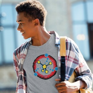 May include: A gray t-shirt featuring a colorful graphic design. The design includes a red circle with a yellow center and blue and yellow connecting shapes. The person is wearing a plaid shirt and carrying a yellow backpack.