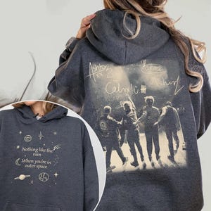 May include: Dark gray hoodie featuring a band photo and signatures on the back. The front displays the text "Nothing like the rain When you're in outer space" with celestial graphics. A comfortable, casual garment.