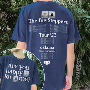 May include: A navy blue t-shirt with white text that reads "The Big Steppers Tour '22 oklama (is not your savior.)" on the back. The front of the shirt has white text that reads "Are you happy for me?" with a small image of a person standing on a beach.