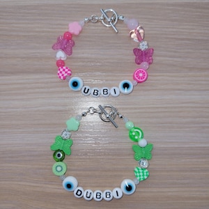 Puede incluir: Dos pulseras de cuentas con cierres plateados. La pulsera superior tiene cuentas rosas, blancas y transparentes con un dije de mariposa, un dije de corazón y un dije de rodaja de limón. La pulsera inferior tiene cuentas verdes, blancas y transparentes con un dije de mariposa, un dije de estrella y un dije de cara sonriente. Ambas pulseras tienen la palabra "UBBI" escrita en letras blancas sobre cuentas negras.