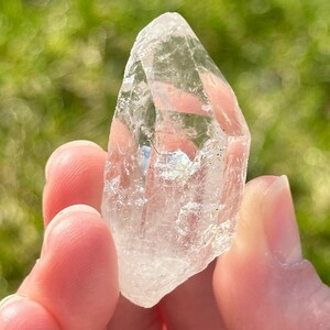 May include: A clear quartz crystal point, held in a person's hand, with a natural, rough texture. The crystal is transparent, allowing light to pass through. The background is blurred green.