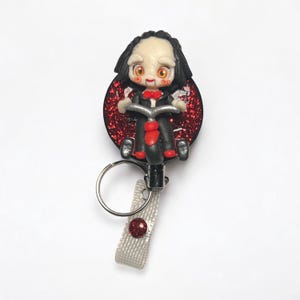 May include: A red glitter badge reel with a black and white clay figure of a character from the movie "Saw" riding a tricycle. The figure has a red bow tie and a creepy smile.