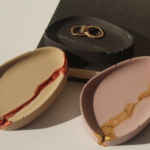 May include: Three oval-shaped concrete trinket dishes with gold and copper accents. The dishes are on a white surface.