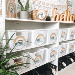 May include: White storage bins with rainbow labels on a white shelf. The labels read "Student Supplies", "Math Games", "Letter Cards", "Glue Sticks", "Dry Erase Markers", "Paper Clips", "Phonics Games", and "Reading Markers".