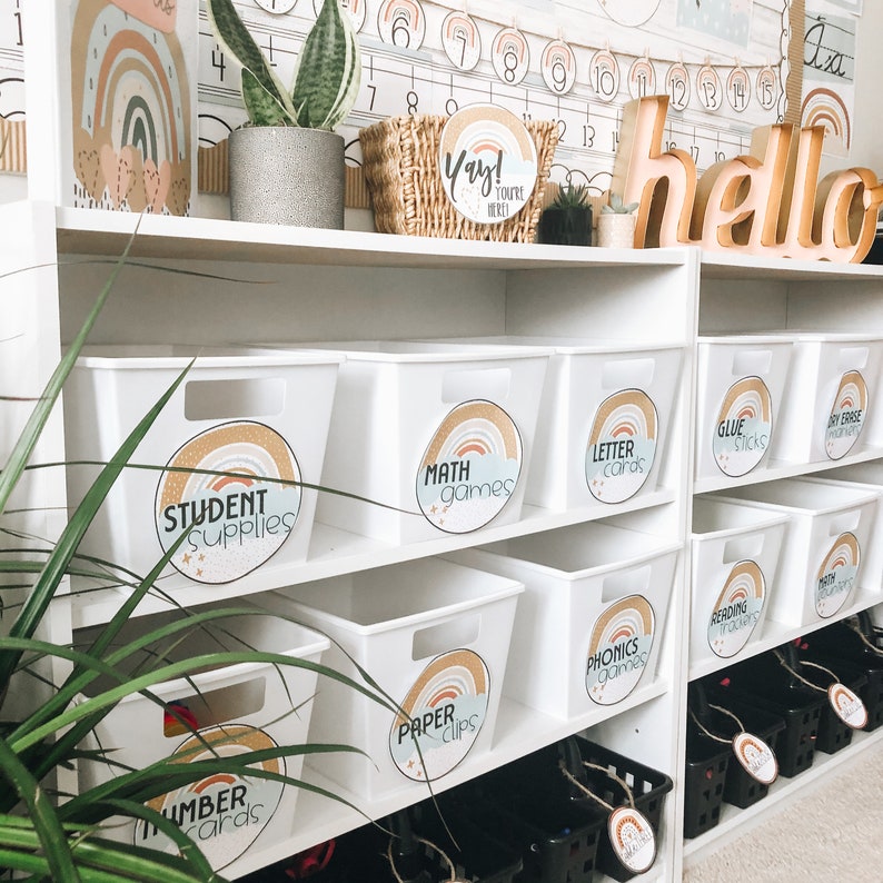 May include: White storage bins with rainbow labels on a white shelf. The labels read "Student Supplies", "Math Games", "Letter Cards", "Glue Sticks", "Dry Erase Markers", "Paper Clips", "Phonics Games", and "Reading Markers".