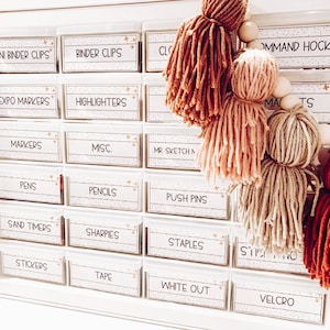 May include: White plastic drawer organizer with labels for various school supplies, including binder clips, highlighters, markers, pens, pencils, push pins, sand timers, sharpies, staples, stickers, tape, white out, and velcro. The drawers are decorated with gold stars. A string of colorful yarn pom-poms hangs in front of the drawers.