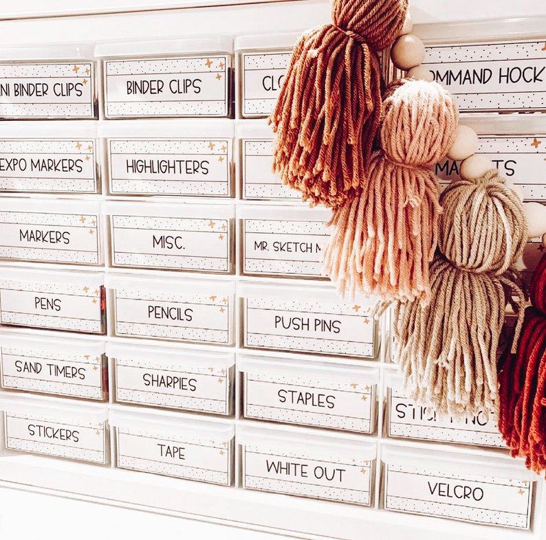 May include: White plastic drawer organizer with labels for various school supplies, including binder clips, highlighters, markers, pens, pencils, push pins, sand timers, sharpies, staples, stickers, tape, white out, and velcro. The drawers are decorated with gold stars. A string of colorful yarn pom-poms hangs in front of the drawers.