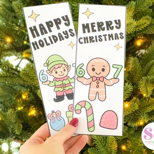 May include: Two Christmas-themed bookmarks are held in front of a Christmas tree. One bookmark says "Happy Holidays" with an elf holding a number 6. The other says "Merry Christmas" with a gingerbread man holding a 6 and a 7.