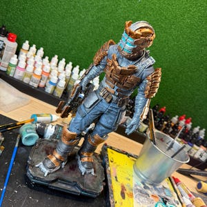 May include: A detailed figurine of a character in a futuristic suit, predominantly blue and bronze. The figure is posed on a textured base, holding a weapon. The helmet has a distinctive design with blue and orange accents. The figure is set against a green backdrop.
