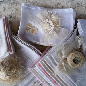 May include: A collection of white and pink-trimmed handkerchiefs, adorned with lace, buttons, and thread. Some handkerchiefs are embellished with lace bows and floral accents, creating a vintage aesthetic. The arrangement suggests a craft or textile theme.