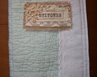 May include: A light blue quilted fabric journal cover with a white lace trim and a burlap tag that says "STITCHES" with a small floral embellishment.