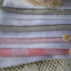 May include: A stack of folded fabric in various shades of white, pink, and gray. The fabrics include linen and other textiles. Two buttons and a bundle of thread are also visible.
