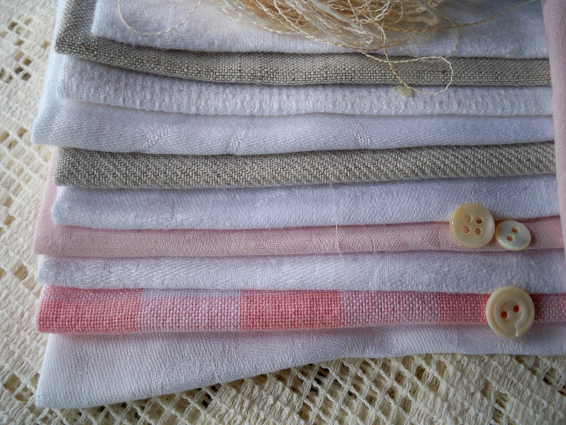 May include: A stack of folded fabric in various shades of white, pink, and gray. The fabrics include linen and other textiles. Two buttons and a bundle of thread are also visible.